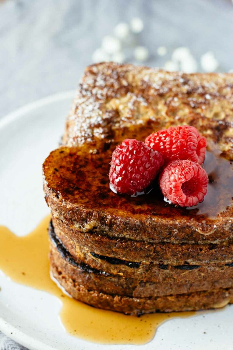 The most delicious Healthy French Toast made with simple ingredients including Ezekiel bread which is packed with healthy grains. This recipe is super easy to whip up and is perfect for lazy weekend breakfast or brunch with a side of eggs and hot cup of coffee. #healthyfrenchtoast #frenchtoast A close up of healthy french toast topped with raspberries on a white plate