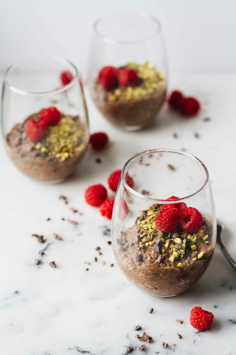 Vegan Chocolate Chia Seed Pudding in a glass cup and topped with raspberries, pistachios, and cacao nibs!