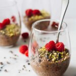 Vegan Chocolate Chia Seed Pudding in a glass cup and topped with raspberries, pistachios, and cacao nibs!