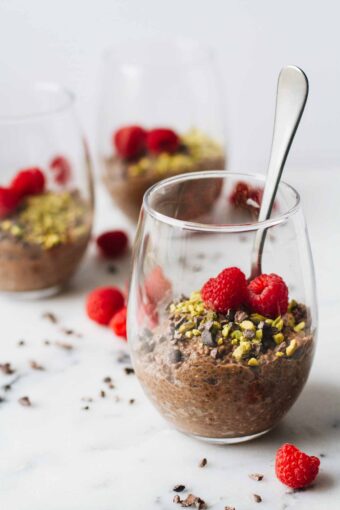 Vegan Chocolate Chia Seed Pudding in a glass cup and topped with raspberries, pistachios, and cacao nibs!