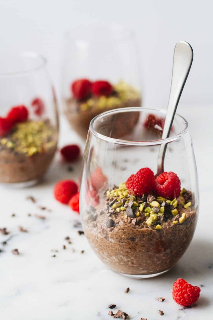Vegan Chocolate Chia Seed Pudding in a glass cup and topped with raspberries, pistachios, and cacao nibs!
