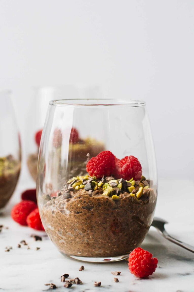 Vegan Chocolate Chia Seed Pudding in a glass cup and topped with raspberries, pistachios, and cacao nibs!