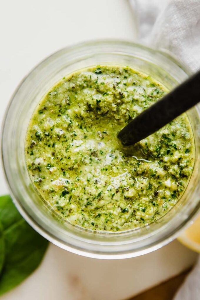 A jar of spinach pesto with a spoon coming out of the top of it.