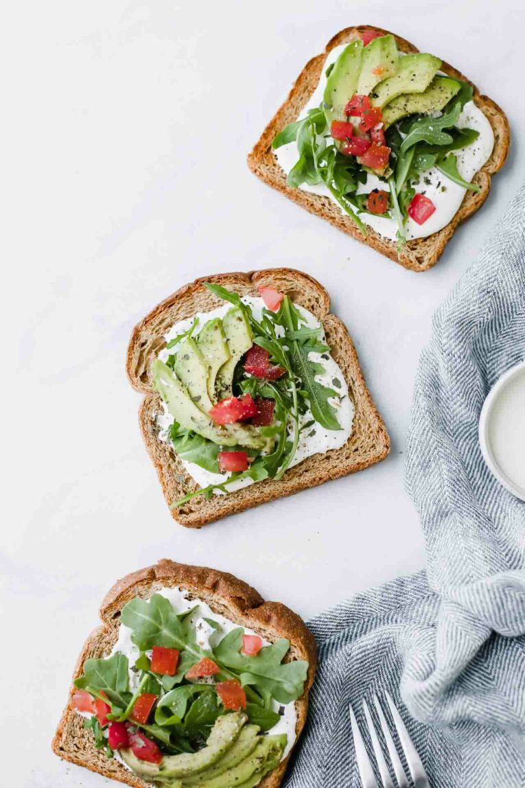 Toasted bread topped with goat cheese, avocado, arugula, and tomatoes over a white marble table.