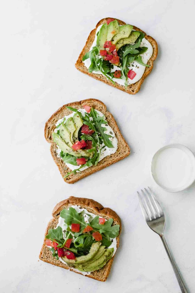 3 pieces of  avocado toast topped with goat cheese, arugula, avocado, and tomatoes on a countertop next to fork. 