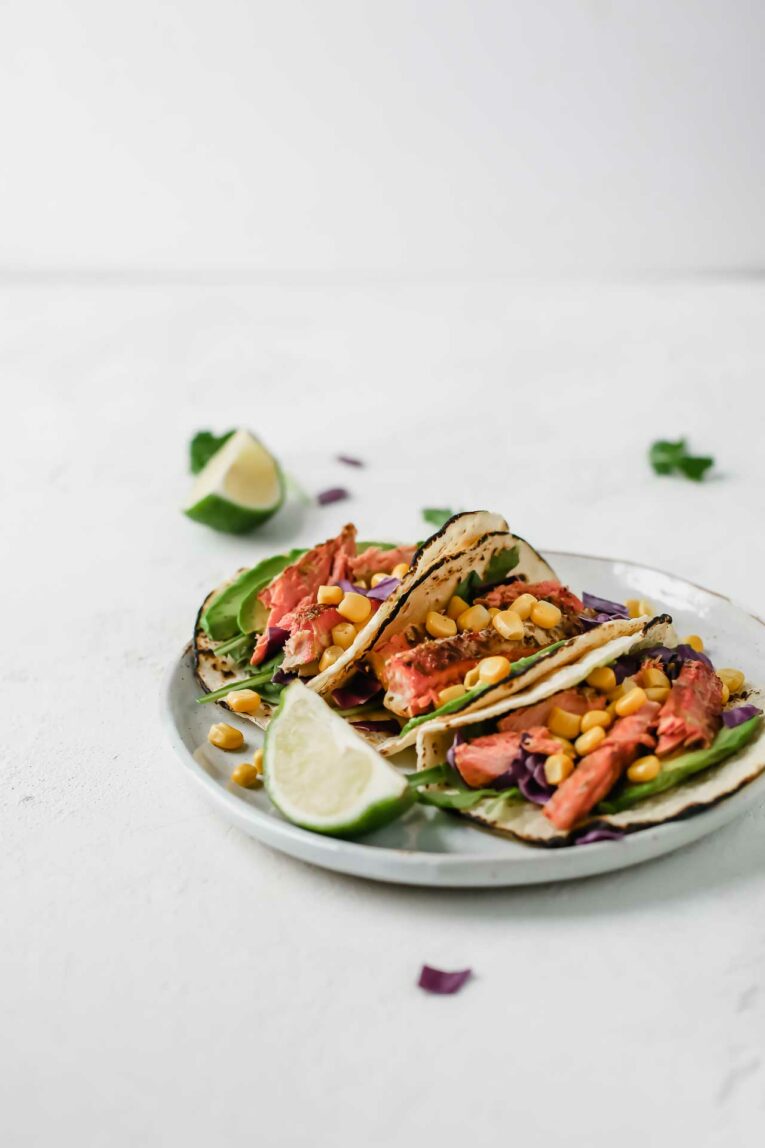 These Spicy Salmon tacos are super easy to put together and taste incredible. Made with juicy salmon fillets, corn, shredded cabbage and drizzled with a tangy and creamy avocado lime dressing. A close up of Spicy Salmon Tacos stuffed with corn and purple cabbage