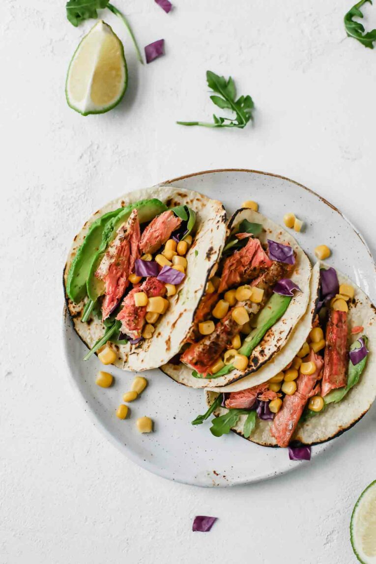 These Spicy Salmon tacos are super easy to put together and taste incredible. Made with juicy salmon fillets, corn, shredded cabbage and drizzled with a tangy and creamy avocado lime dressing. #jaroflemons #salmontacos #tacos Spicy Salmon Tacos on a grey plate topped with avocado slices
