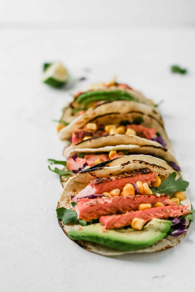 These Spicy Salmon tacos are super easy to put together and taste incredible. Made with juicy salmon fillets, corn, shredded cabbage and drizzled with a tangy and creamy avocado lime dressing. A close up of Spicy Salmon Tacos on a grey table
