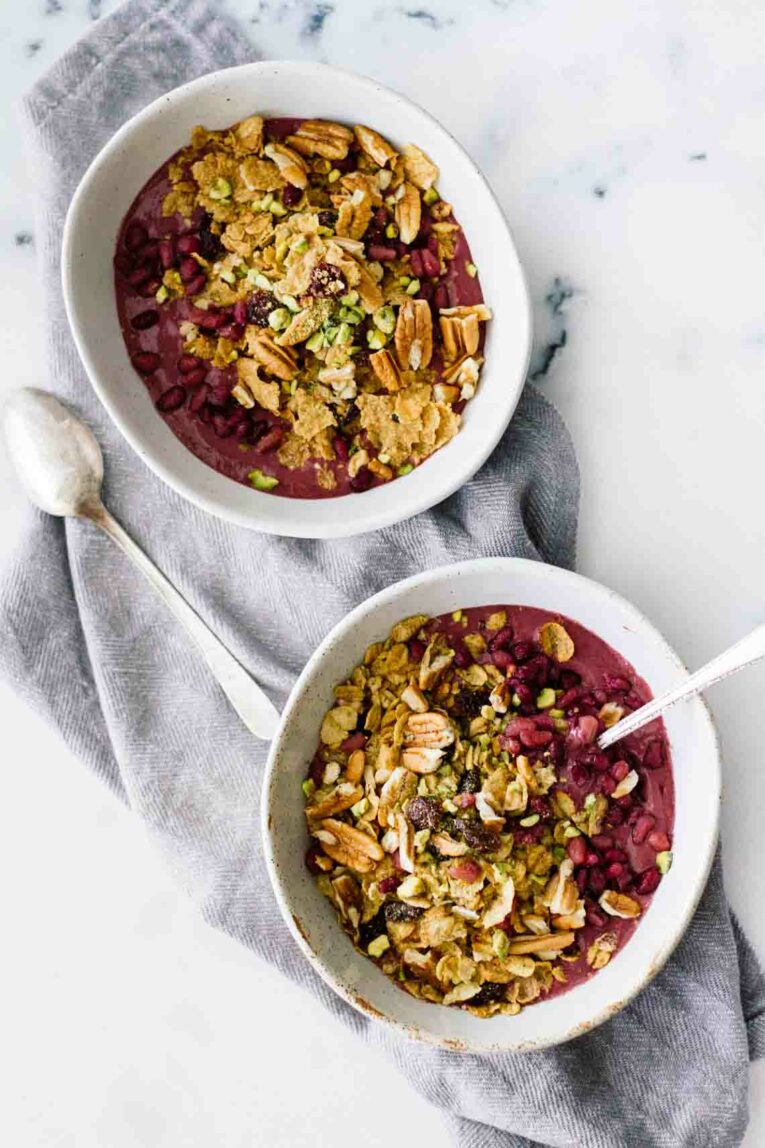 An overhead shot of two acai bowls topped with fruit and nuts sitting on a marble surface