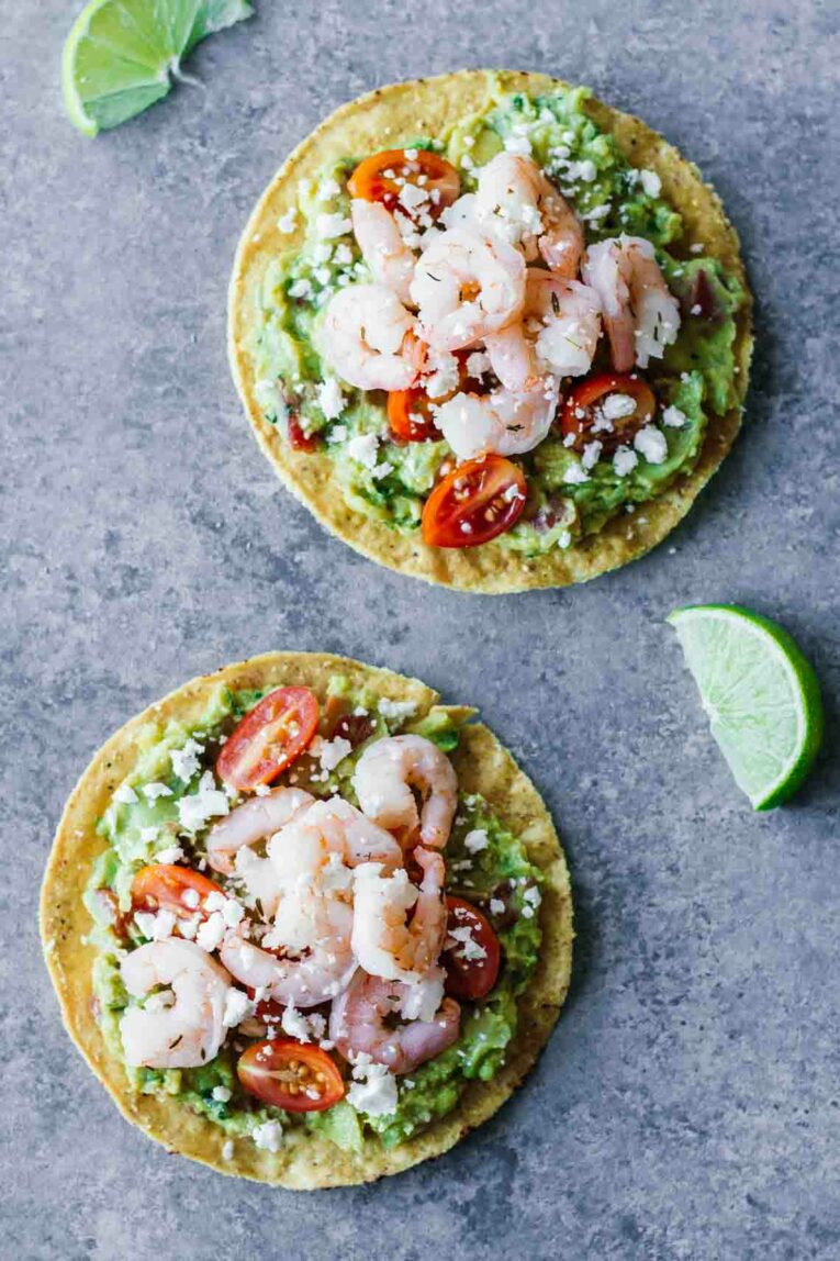 overhead shot of twoShrimp Guacamole Tostadas on grey surface next to cut limes