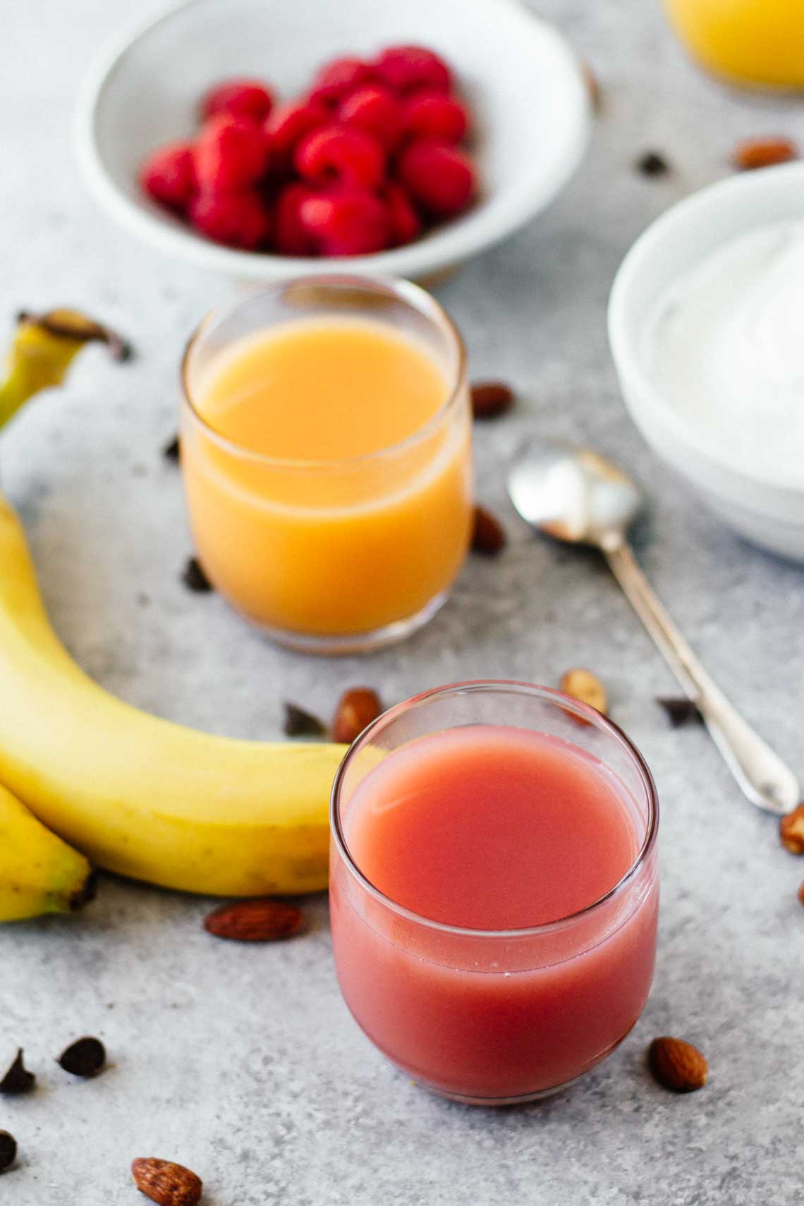 two different juices on a counter with a bowl of raspberries and bananas in the background