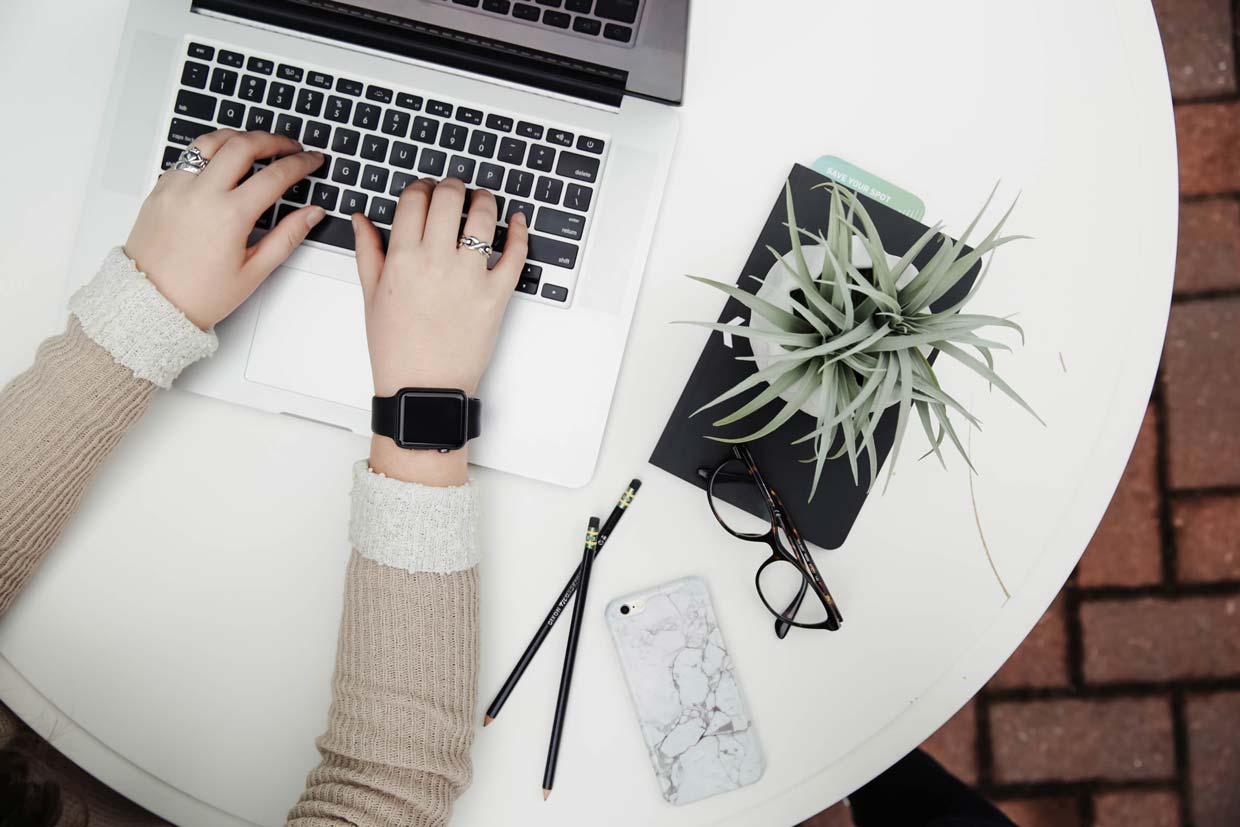  Hands Typing on a computer next to a plant, glasses, pencils, and a phone.