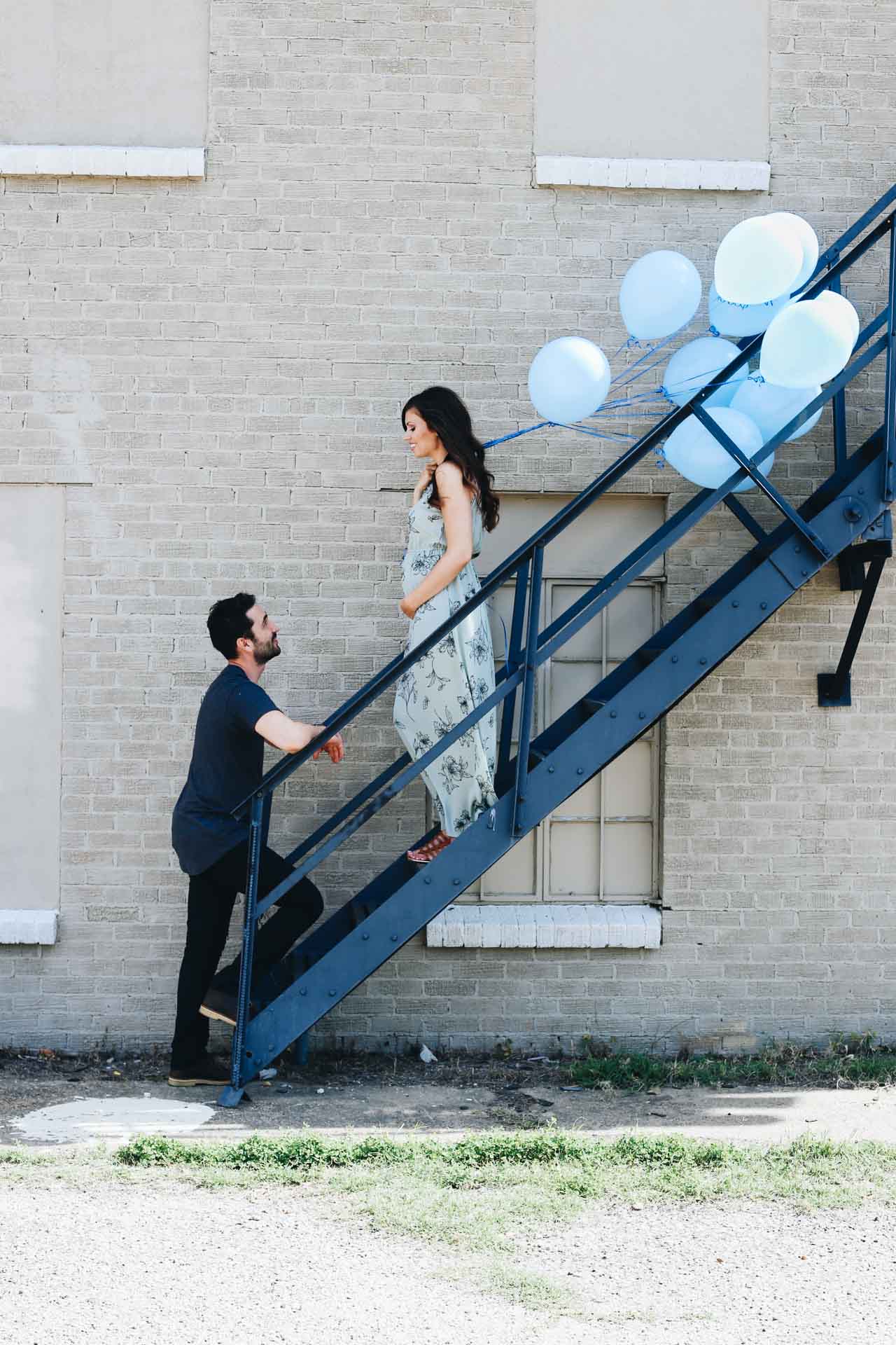 Couple standing on metal stairs outside. Girl holding blue balloons. 