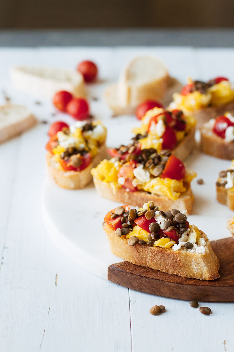 Close up of Tomato & Lentil Breakfast Bruschetta scattered on a wooden surface.