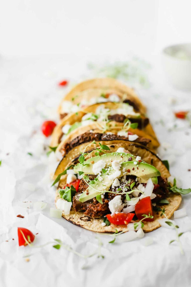 Side angle of  Corn tortillas topped with slow cooked chuck roast beef, avocado, tomatoes, onions, and feta cheese on a white surface.