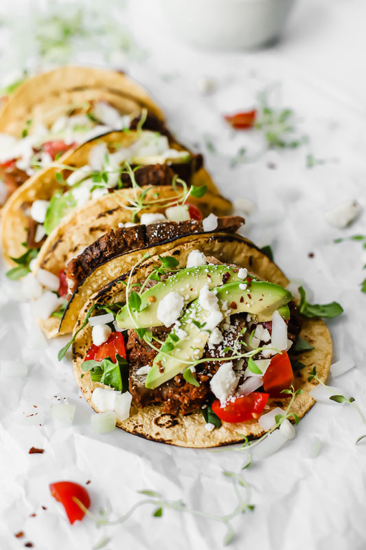  Corn tortillas topped with slow cooked chuck roast beef, avocado, tomatoes, onions, and feta cheese on a white surface.