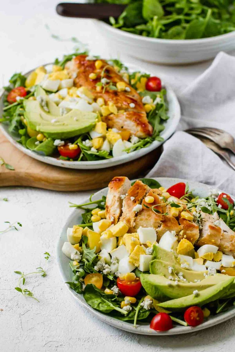 Healthy Chicken Cobb Salad Recipe loaded with spicy greens, creamy avocado, boiled eggs, fresh tomatoes, corn, delicious grilled chicken, and topped with a sprinkle of blue cheese crumbles served on two white plates.