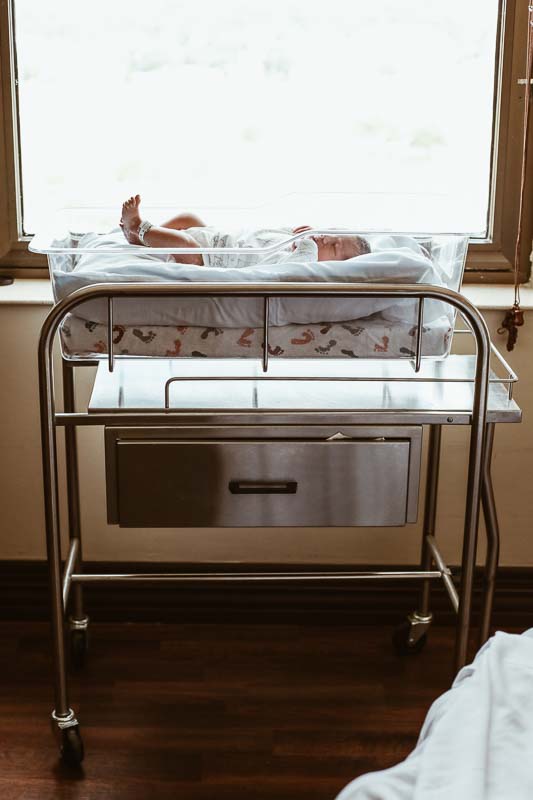 newborn baby sitting in hospital bassinet next to window.