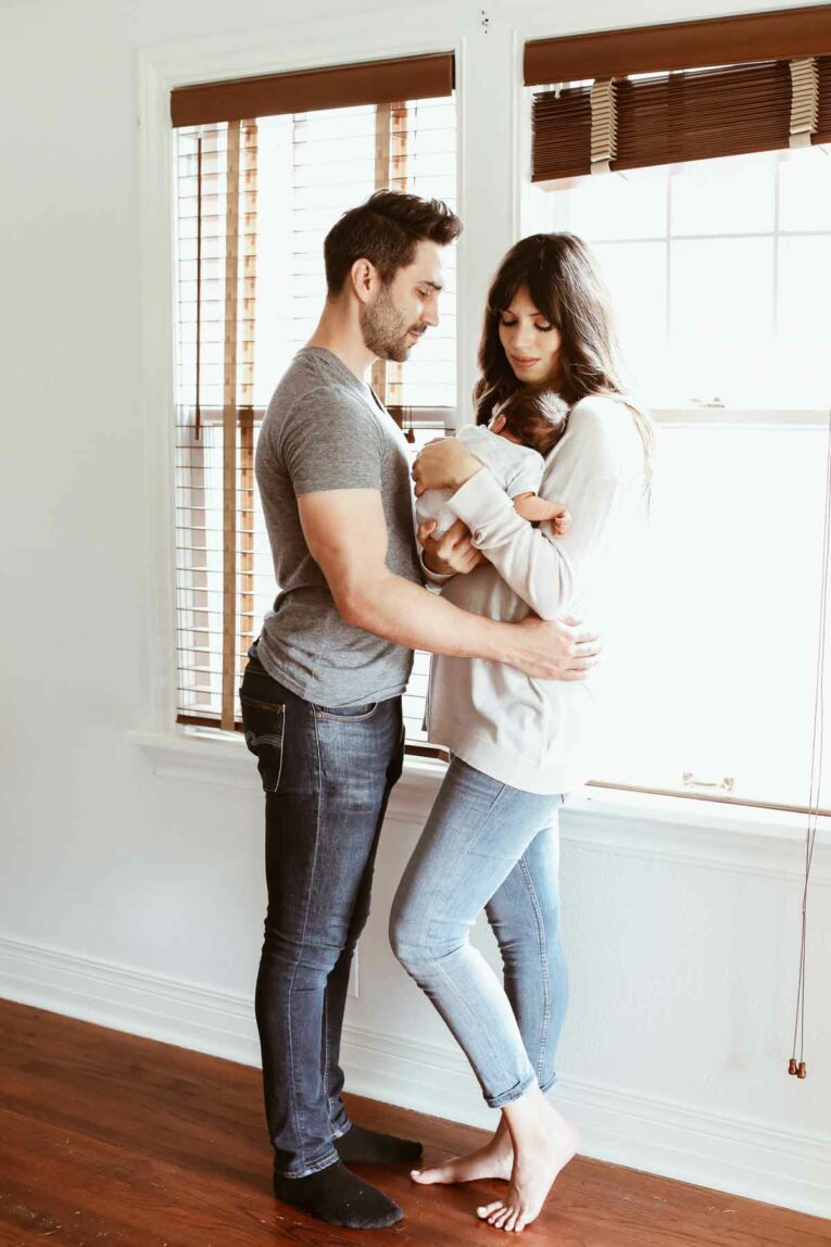 Couple standing next to windows holding newborn baby.