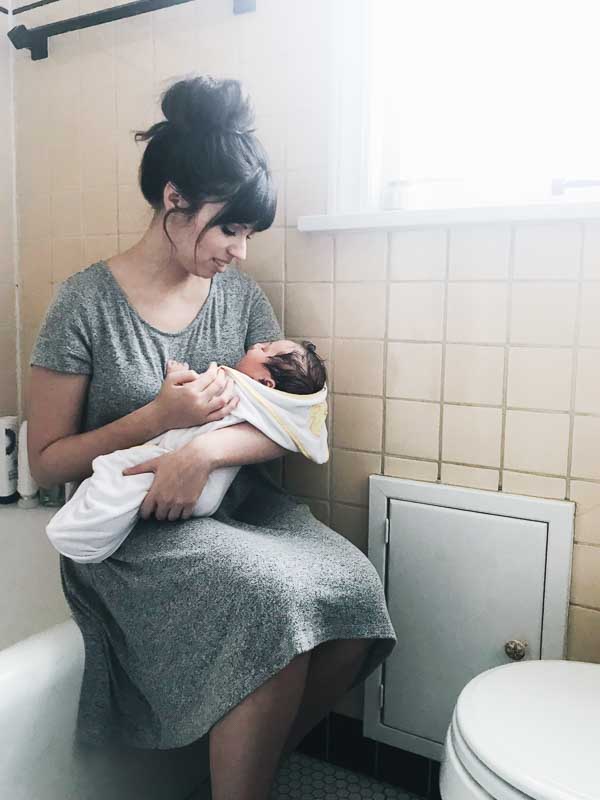 Mom sitting on bathtub holding freshly bathed baby next to window.