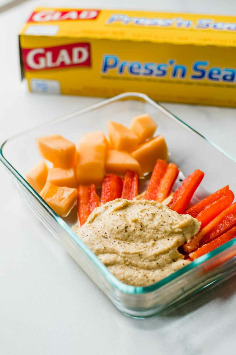side angle of carrots, mango, and hummus in a glass container next to glad press'n seal box.