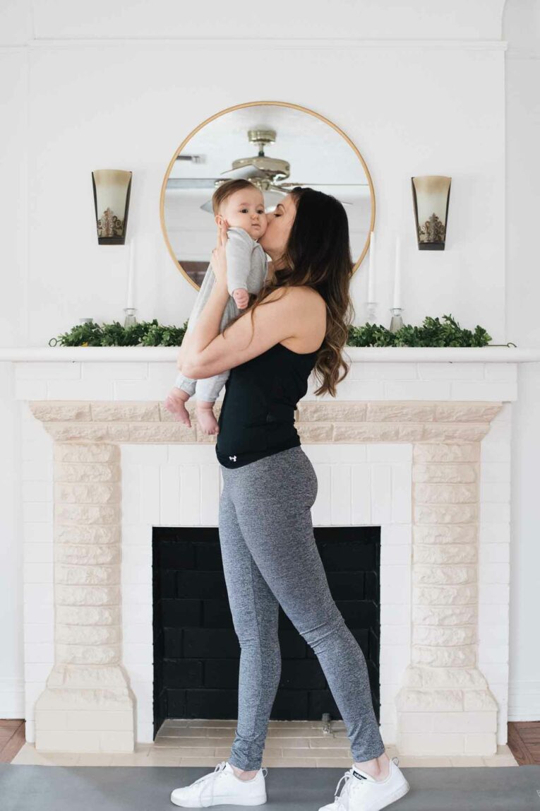 Woman wearing fitness clothes and kissing baby while standing on yoga mat