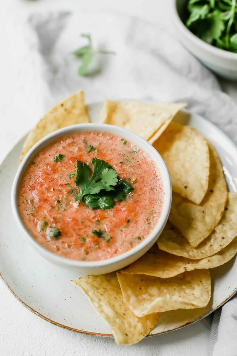 The BEST Easy Salsa Recipe made with plum tomatoes, jalapeño, and cilantro. So yummy! Angled close up of the best easy salsa recipe in a white bowl on top of a grey plate surrounded with tortilla chips