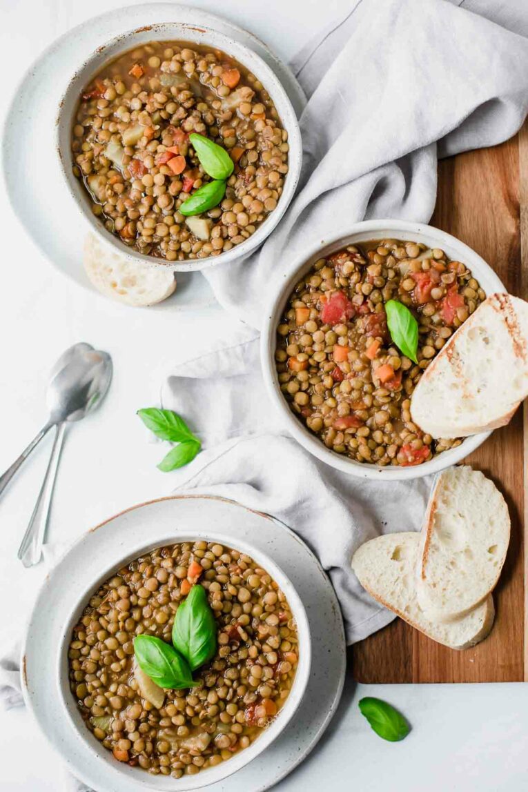 Three bowls of vegan lentil soup served with fresh basil and toasted baguette slices.