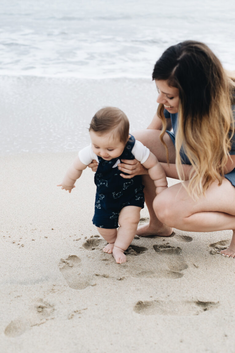 Heading to the beach with your baby? Here are the best beach essentials you will need to take with you for your baby (and some great things for mom, too)! Baby walking on the beach while mother holds him.