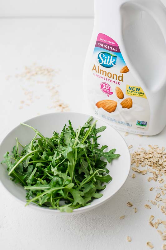 Bowl of arugula on a table with oats and almond milk.
