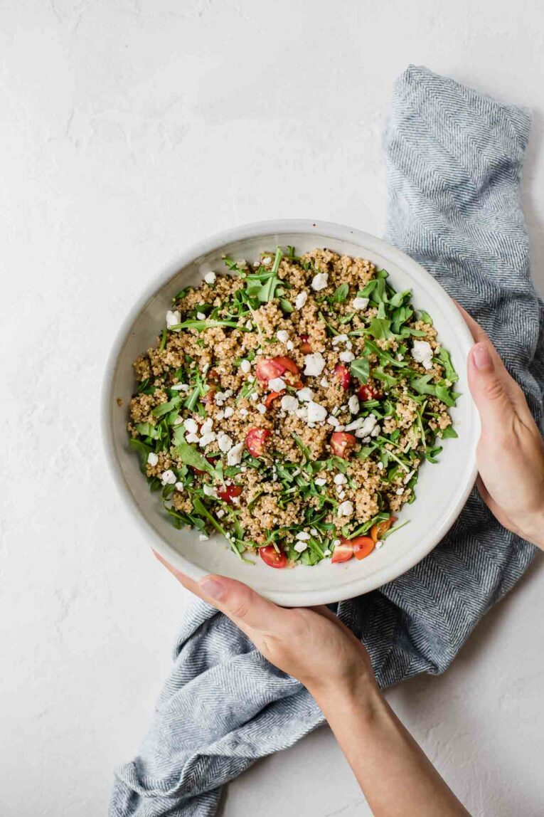 Hands holding white bowl of quinoa salad topped with arugula, tomatoes, and feta cheese.