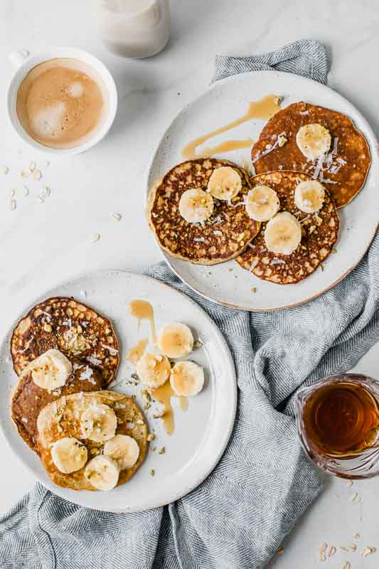 The BEST ever Healthy Banana Pancakes that can be made in 10 minutes or less! Made with real banana, protein-loaded eggs, rolled oats, and plant-based protein powder. So delicious and perfect for your entire family! Two plates of healthy banana pancakes on a table with coffee and syrup.