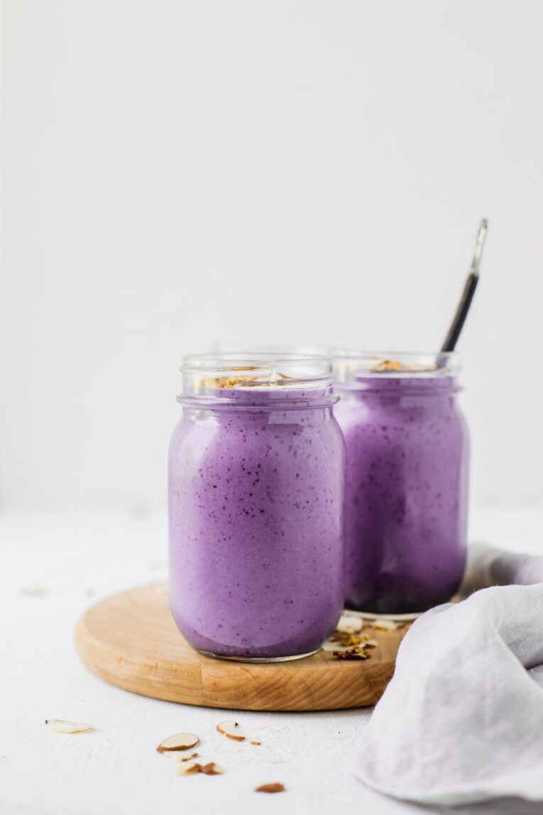 Creamy, sweet, tasty, and one of the BEST protein shakes out there! This Blueberry Vanilla Protein Shake Recipe is made with fresh or frozen blueberries, creamy greek yogurt, your choice of milk, and vanilla protein powder. Make this easy snack or breakfast in 5 minutes or less! Blueberry Vanilla Protein Shakes on a wood cutting board.