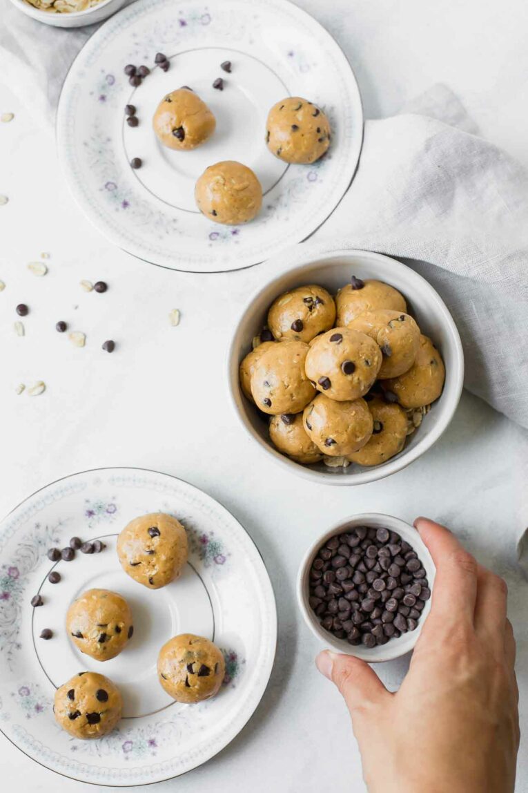 The BEST Peanut Butter Energy Balls that only take about 10 minutes to make. Keep these healthy bites on hand for easy snacking or take them on-the-go! (Vegan, vegetarian, gluten-free, dairy-free, refined sugar free) Vegan peanut butter energy balls on plates and a bowl next to chocolate chips.