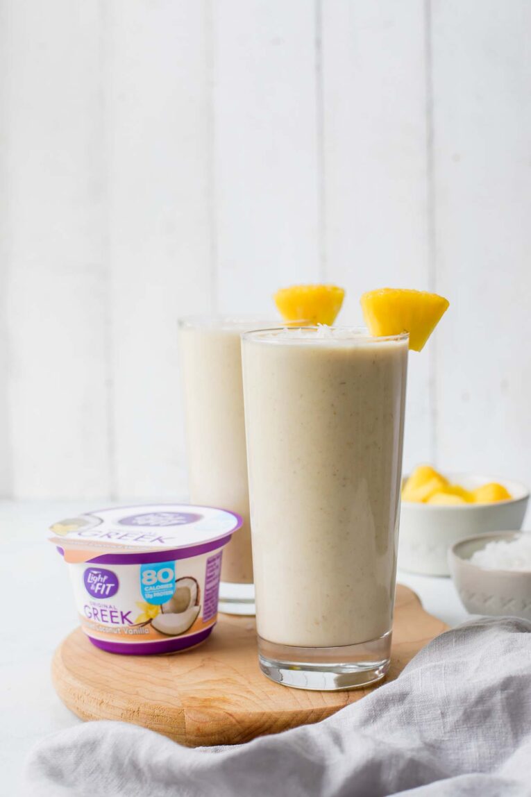 pineapple coconut vanilla smoothie served in two glasses on a cutting board next to a container of yogurt.