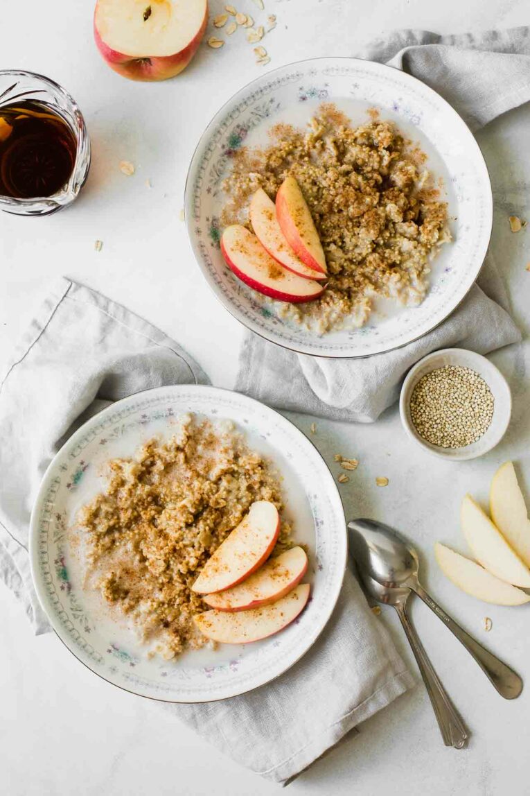 Sweet, wholesome, delicious, and easy to make. We're loving this simple Apple Cinnamon Quinoa Breakfast Bowl for a healthy start to the day! Quinoa breakfast bowls with apples, maple syrup, and cinnamon on a white marble countertop.