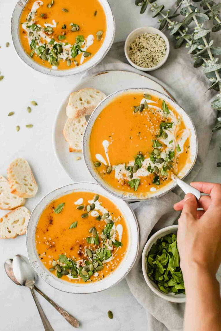 3 bowls of sweet potato soup with a hand holding a spoon.