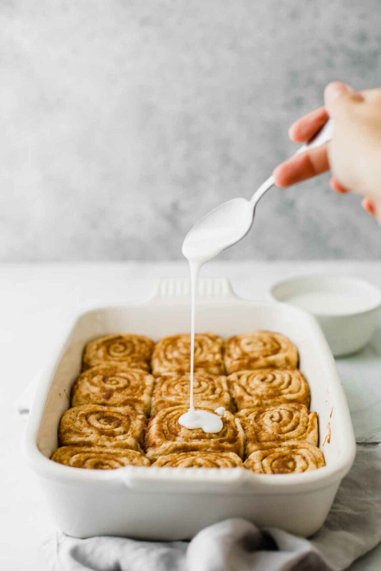 Hand pouring royal icing over baked vegan cinnamon rolls in a white casserole dish.