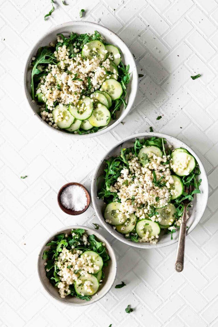 3 white salad bowls with spinach, cucumbers, and quinoa on a white table.