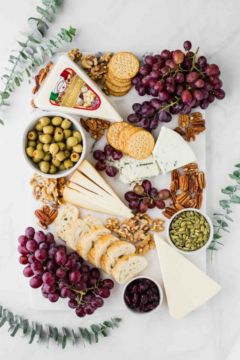 Marble cheese board with 4 wedges, baguette, crackers, pecans, walnuts, olives, and grapes.