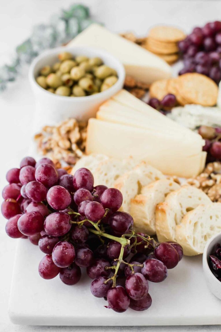 Angled shot of grapes, baguette, cheese wedge, olives, pecans, and crackers on a marble cheese board.