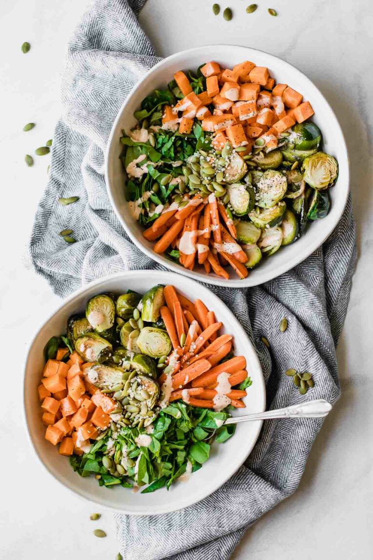Two bowls of spinach, carrots, sweet potatoes, brussels sprouts, and pepitas topped with a creamy sauce.