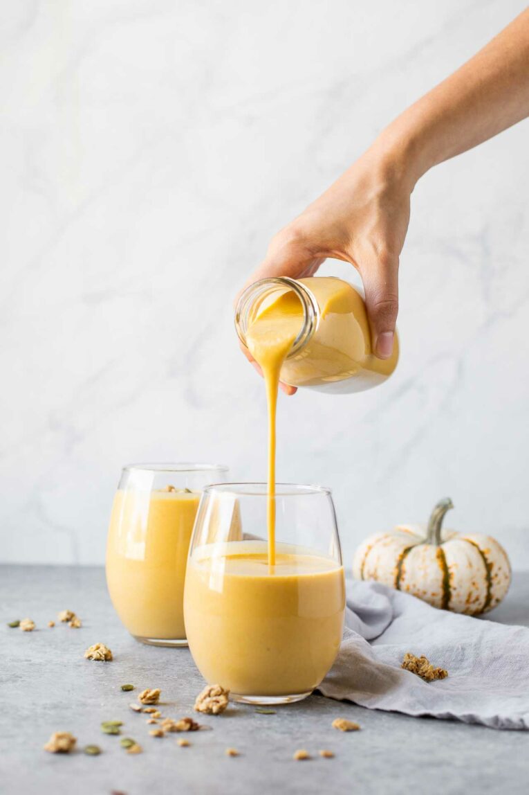 Hand pouring pumpkin smoothie from glass milk jug into glass cup.