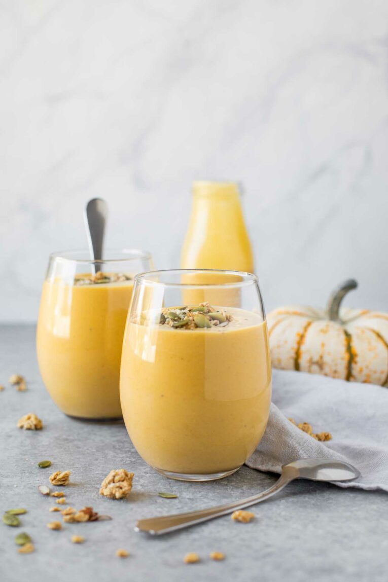 Two Pumpkin Smoothies in a glass with spoons, milk jug, and pumpkin in the background.