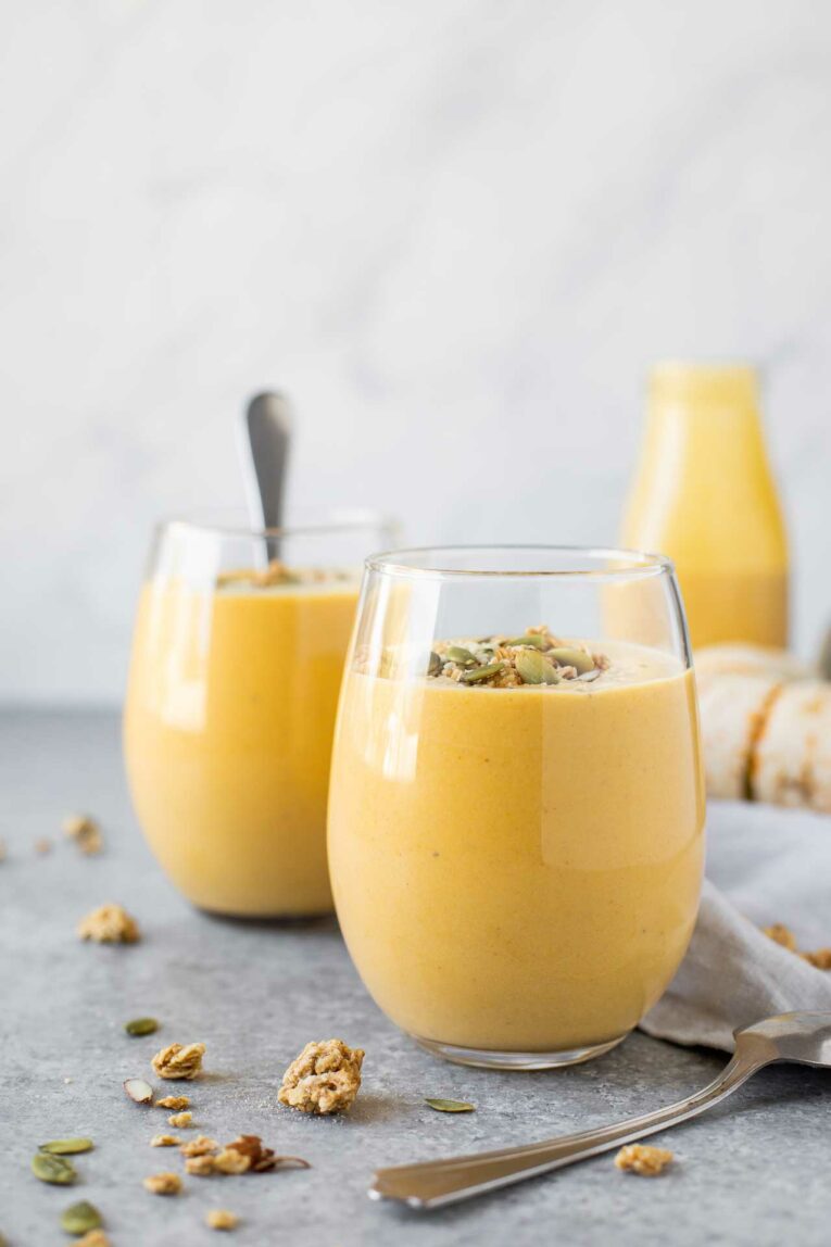 Three pumpkin smoothies in glass cups on a grey table with granola and spoons.
