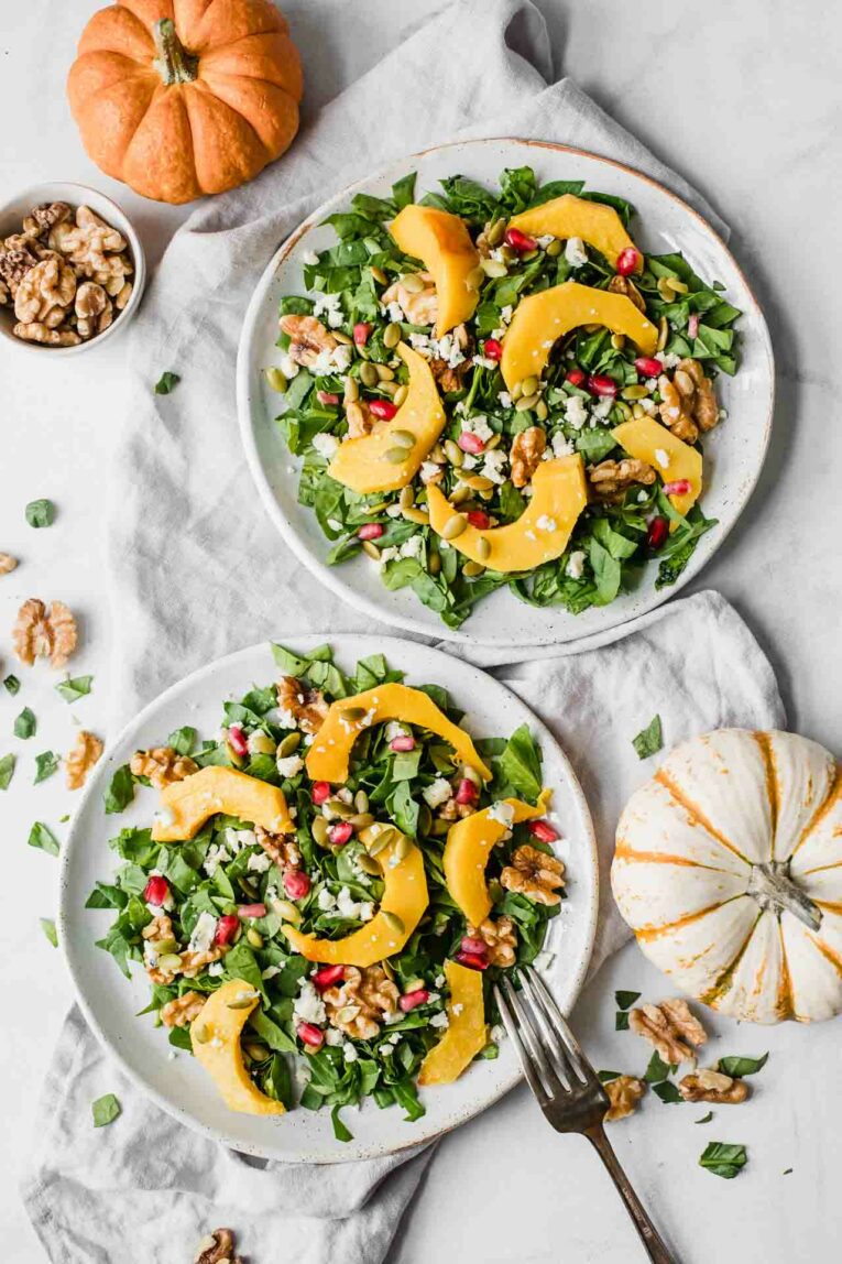 Two plates of roasted pumpkin salad next to pumpkins and a bowl of walnuts.
