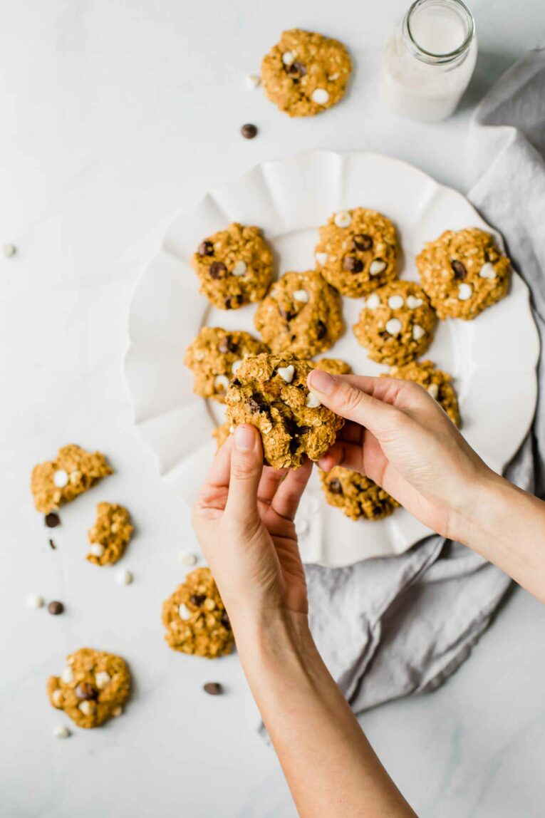 Hands breaking a soft pumpkin oatmeal cookie in half over a white plate.