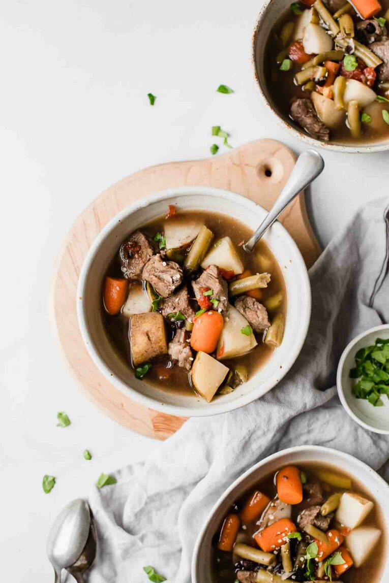 Top shot of 3 white bowls of beef stew with spoons and chopped spinach.