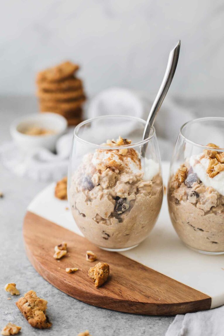 Angled shot of two glasses of oatmeal cookie overnight oats with a spoon on a cutting board.