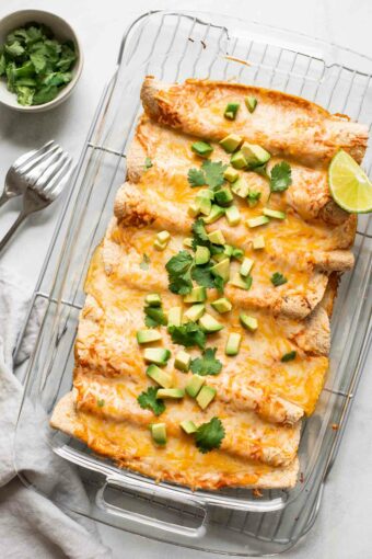 Enchiladas in a casserole dish topped with cheese, cilantro, avocado, and lime.